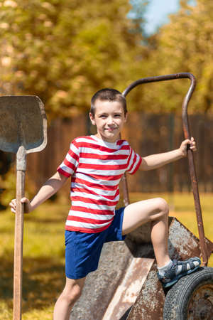 A little cheerful boy sits on a garden wheelbarrow and holds a shovel in his hand in the garden of a country house. Little boy helper ready to digの写真素材