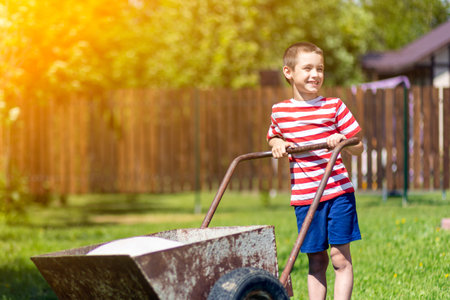 A little boy drives a wheelbarrow with bags on a sunny day in the garden. Boy assistant in the gardenの写真素材