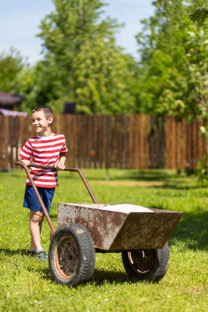 A little boy drives a wheelbarrow with bags on a sunny day in the garden. Boy assistant in the gardenの写真素材