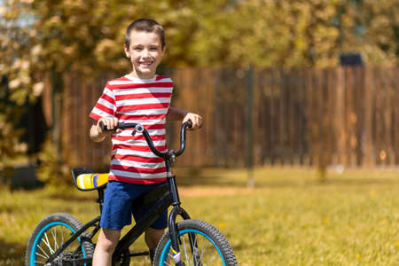 Happy cheerful boy in a striped t-shirt is doing well, looking at the camera and sitting on a bike against the background of the garden. Active outdoor games for children in summer.の写真素材