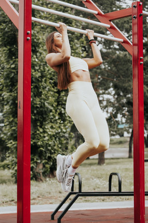 Young woman with long straight hair on street workout pulls up on a barbell on the sports ground in...