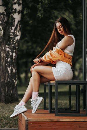Young beautiful brunette woman in a short white dress posing on a wooden bench against the background of a green city parkの写真素材