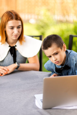 Mom and boy schoolboy are engaged in lessons through a laptop at home in the garden. Online classes for children. Schoolboy listens to a lecture and solves problemsの写真素材