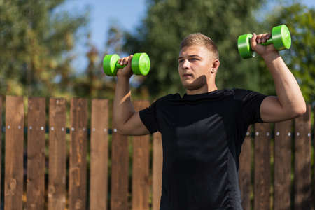 A young man goes in for sports. The athlete does morning exercises with dumbells, bends and pulls his hand in garden insummer dayの写真素材