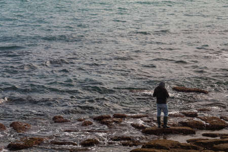 a male fisherman with a black raincoat catches fish on the sea in the winter seasonの写真素材