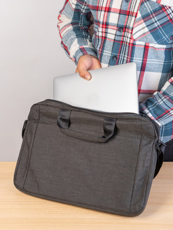 A man puts a laptop in a bags outside, on a light background. A young man packs a laptop into a backpack. A man pulls a laptop out of his backpack.の写真素材