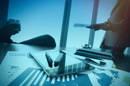 business documents on office table with smart phone and digital tablet and stylus and two colleagues discussing data in the background with twilight effectの写真素材