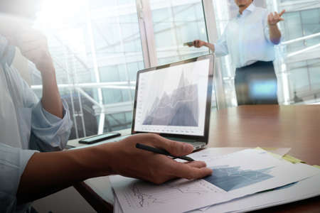 business documents on office table with smart phone and laptop computer and graph business with social network diagram and two colleagues discussing data in the backgroundの写真素材