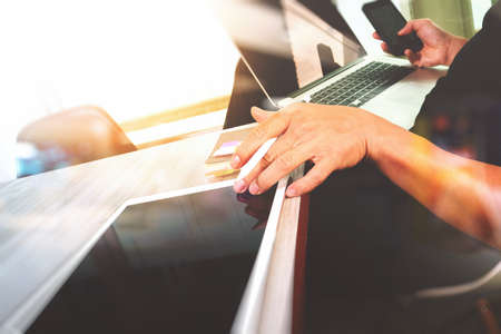 designer hand working and smart phone and blank screen laptop and digital tablet on wooden desk in officeの写真素材
