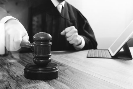 justice and law concept.Male judge in a courtroom striking the gavel,working with digital tablet computer docking keyboard on wood table,black and whiteの写真素材
