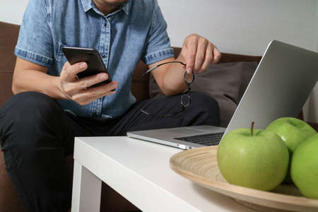 hipster hand using laptop compter and mobile payments online business,eyeglasses,sitting on sofa in living room,green apples in wooden trayの写真素材