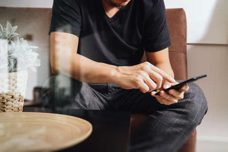 designer man hand using smart phon for mobile payments online shopping,omni channel,sitting on sofa in living room,vase rattan with plant and wooden tray on table,filterの写真素材