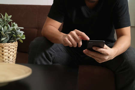 designer man hand using smart phon for mobile payments online shopping,omni channel,sitting on sofa in living room,vase rattan with plant and wooden tray on tableの写真素材
