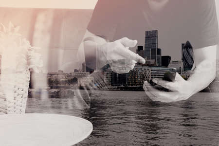 double exposure of designer man hand using smart phon for mobile payments online shopping,omni channel,sitting on sofa in living room,vase rattan with plant and wooden tray on table,London architecture cityの写真素材