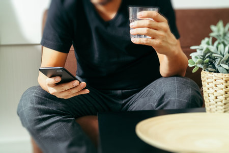 designer man hand with glass of water using smart phon for mobile payments online shopping,omni channel,sitting on sofa in living room,vase rattan with plant and wooden tray on tableの写真素材