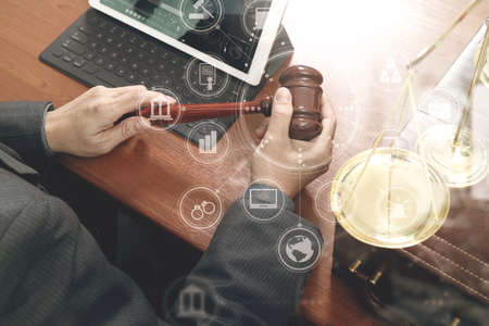 justice and law concept.Male judge in a courtroom with the gavel,working with smart phone,digital tablet computer docking keyboard,brass scale,on wood table,virtual interface graphic icons diagramの写真素材