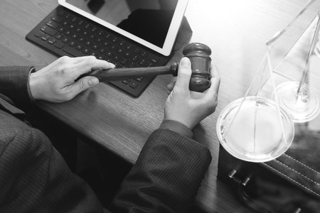 justice and law concept.Male judge in a courtroom with the gavel,working with smart phone,digital tablet computer docking keyboard,brass scale,on wood table,black and whiteの写真素材