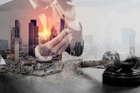 double exposure of justice and law concept.Male judge in a courtroom with the gavel,working with smart phone,digital tablet computer docking keyboard,brass scale,on wood table,London architecture cityの写真素材