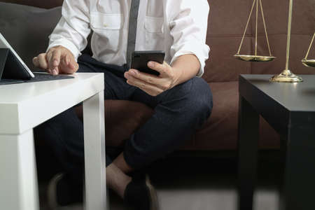 Justice and Law context.Male lawyer hand sitting on sofa and working with smart phone,digital tablet computer docking keyboard with gavel and document on living table at homeの写真素材