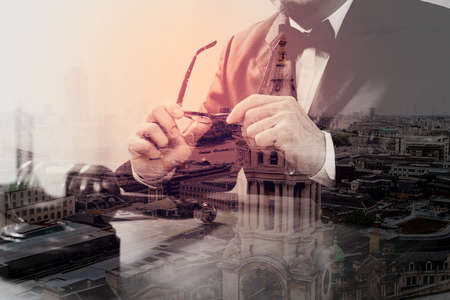 double exposure of justice and law concept.Male judge in a courtroom with the gavel,working with smart phone,digital tablet computer docking keyboard,eyeglasses,on wood table,London architecture cityの写真素材