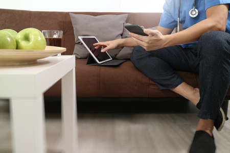 Medical and Health context,doctor hand working with smart phone,digital tablet computer,stethoscope,sitting on sofa in living roomの写真素材