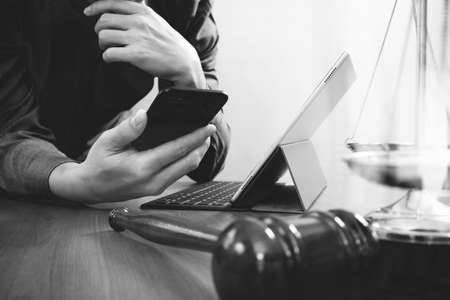 justice and law concept.Male lawyer in office with the gavel,working with smart phone and digital tablet computer and brass scale on wood table,black and whiteの写真素材
