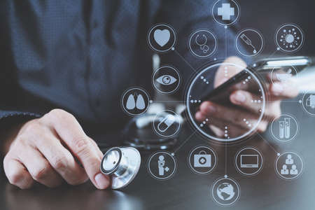 close up of smart medical doctor working with smart phone and stethoscope on dark wooden desk with virtual icon diagramの写真素材