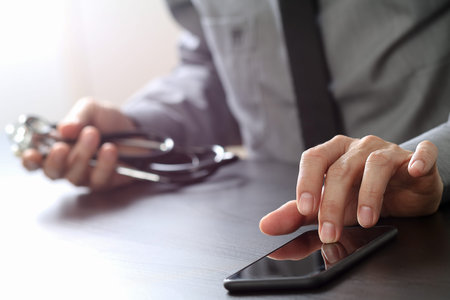 close up of smart medical doctor working with mobile phone and stethoscope on dark wooden deskの写真素材