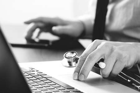 close up of smart medical doctor working with laptop computer and mobile phone and stethoscope on dark wooden desk,black and whiteの写真素材