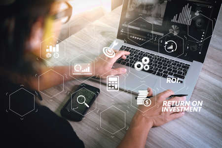 ROI Return on Investment indicator in virtual dashboard for improving business. designer hand attending video conference with laptop computer and smart phone at desk in officeの写真素材