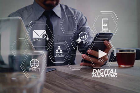 Digital marketing media (website ad, email, social network, SEO, video, mobile app) in virtual screen.success businessman hand using smart phone,digital tablet docking smart keyboard,coffee cup on wooden deskの写真素材
