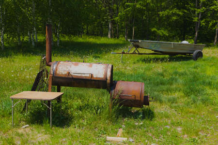 Old round metal wood-burning stove with a chimney, background summer birch forest meadow with dandelions metal motor boat on a trailer.の写真素材