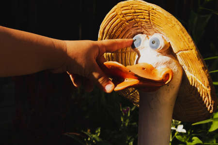 A child's hand, a child's index finger shows the eye of a statuette of a goose in a straw hat in a summer garden.の写真素材