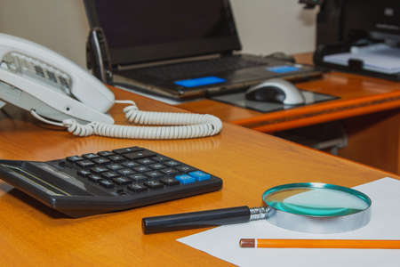 Desktop wooden with a magnifying glass a calculator a phone a pencil a sheet, the background blurred laptop.の写真素材