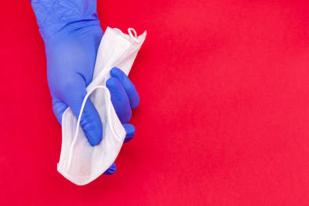 Medical mask, covid-19, a hand in a blue medical disposable glove clutches a white mask on a red background, the concept of the end of the coronavirus.の写真素材