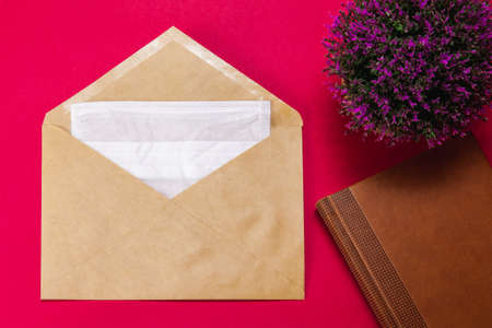Medical face mask in a gray envelope on a red background with a brown Notepad and a lilac flower, close up.の写真素材
