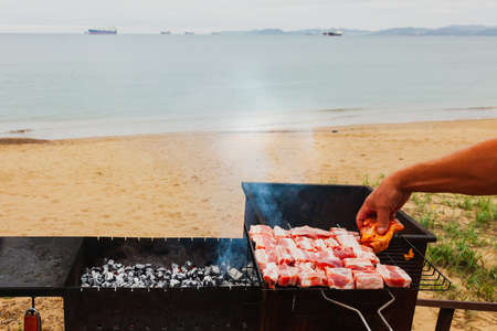 Grilled meat, hand lays pork on the grill over the coals on the background of the sea coast and ships, relaxing on the beach.の写真素材