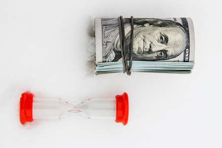 A dollar in winter, a roll of rolled dollar bills lying on the snow with a red hourglass, close-up, the concept of time making money.の写真素材
