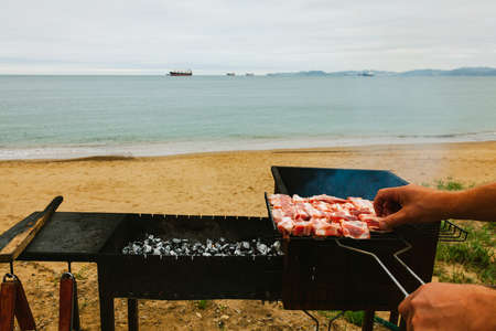 Grilled meat, hand lays pork on the grill over the coals on the background of the sea coast and ships, relaxing on the beach.の写真素材