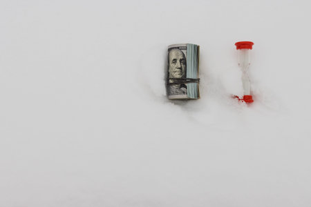 A dollar in winter, a roll of rolled dollar bills lying on the snow with a red hourglass, close-up, the concept of time making money.の写真素材