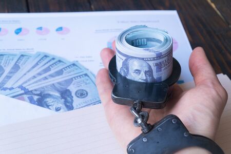 mans handcuffed hand holds twisted money against a background of hundred dollar bills in an envelopeの写真素材