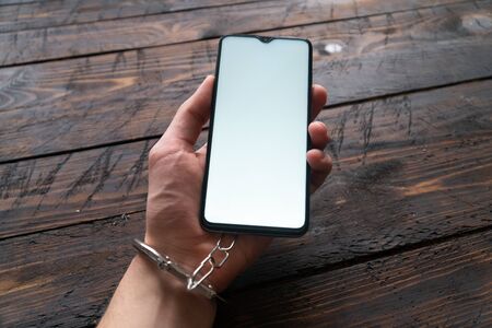 Top view men hand in handcuffs hold modern smartphone with blank screen on wooden brown background. The concept of internet and gadget dependency.の写真素材