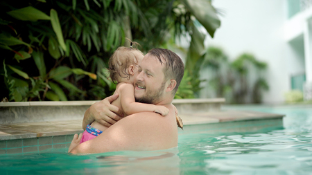 Cute little kid and his father having a swimming lesson in the pool. Father holds daughter in his arms and hugsの写真素材