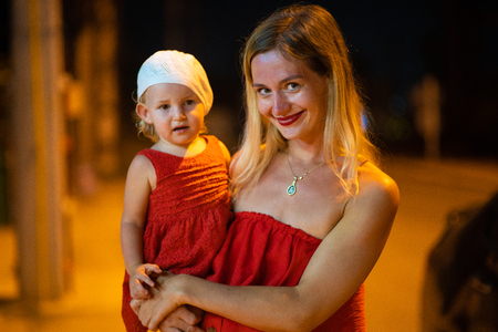 mother holds daughter in her arms, mother with daughter is dressed in red dresses, child in white beret, at nightの写真素材