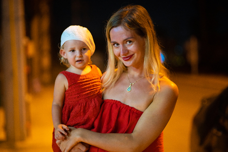 mother holds daughter in her arms, mother with daughter is dressed in red dresses, child in white beret, at nightの写真素材