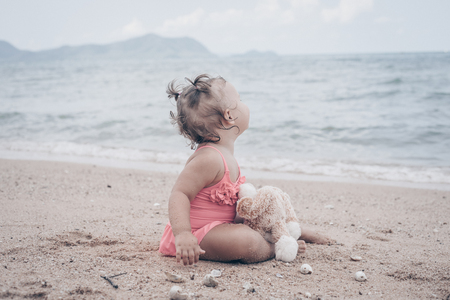 pensive little girl hugging teddy bear and looking away while sitting on seashoreの写真素材