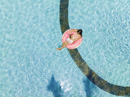 Aerial view of a young brunette woman swimming on an inflatable big donut with a laptop in a transparent turquoise poolの写真素材