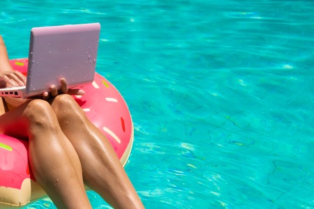 Aerial view of a young brunette woman swimming on an inflatable big donut with a laptop in a transparent turquoise pool, close up.の写真素材