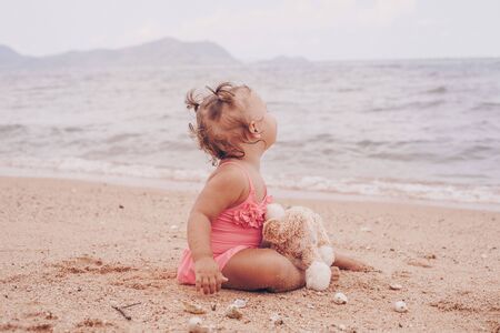 pensive little girl hugging teddy bear and looking away while sitting on seashoreの写真素材