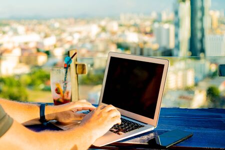 male typing on a laptop in a cafe on the roof of a high-rise with a beautiful panoramic view of the city, close up.の写真素材